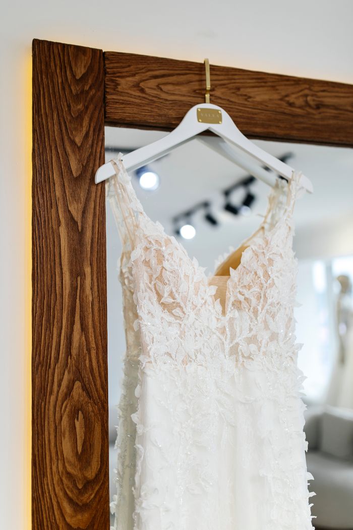A detailed lace wedding dress elegantly hung on a wooden framed mirror in a bright bridal boutique.