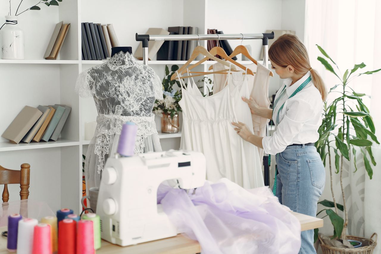 A fashion designer arranges dresses on hangers in a bright, creative studio with sewing tools and mannequins.